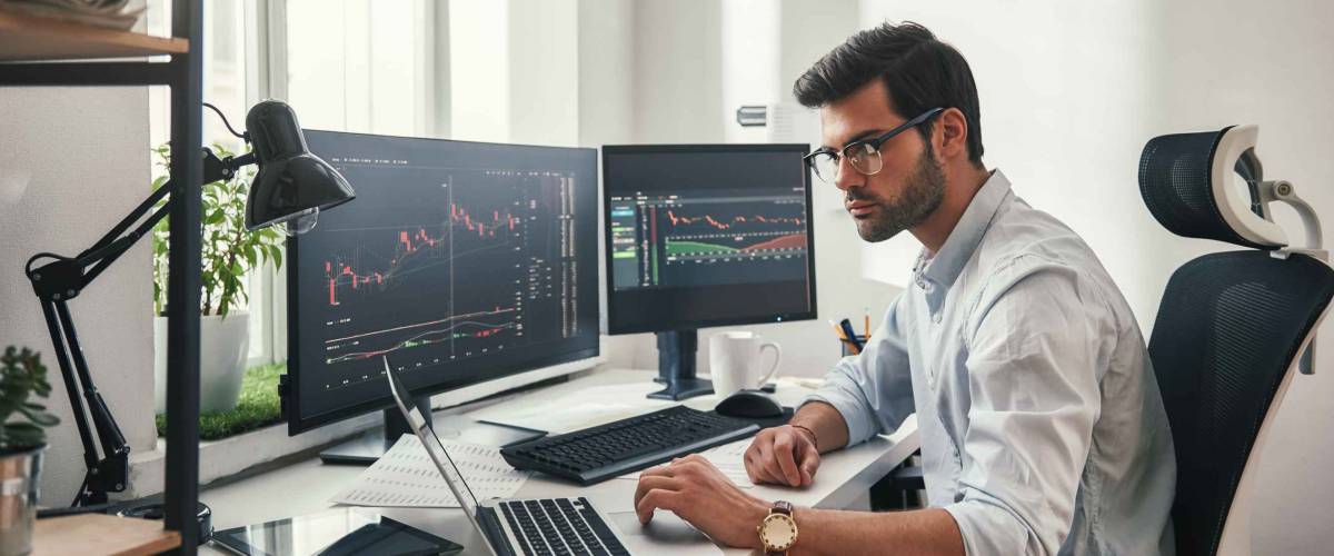 Busy working day. Young bearded trader in eyeglasses working with laptop while sitting in his modern office in front of computer screens with trading charts.