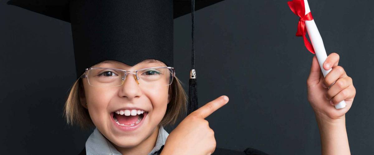 Close-up portrait of happy girl in mortar board holding diploma at the black chalkboard in classroom