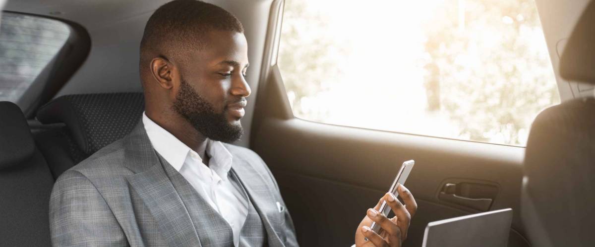 Handsome african american businessman with laptop holding smartphone while going by car on business trip, copy space. Black entrepreneur looking at mobile phone screen, using laptop