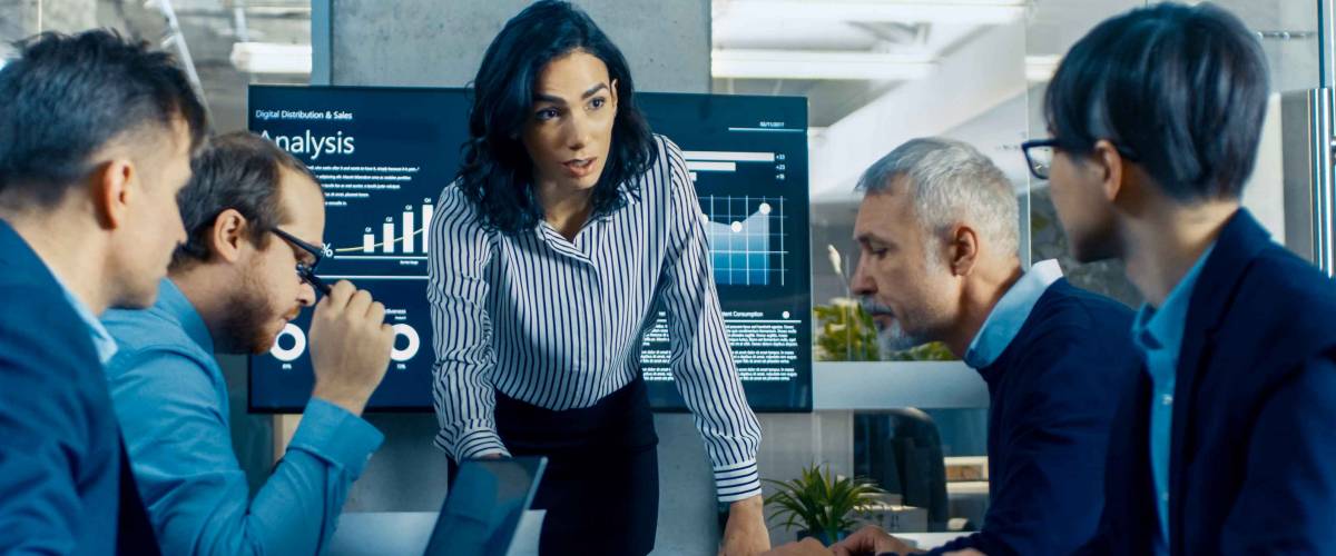Chief Female Executive Leans and Spreads Project Blueprints on the Table Showing them to Her Colleagues. In the Background Pie Charts and Company's Growth on the Wall TV.
