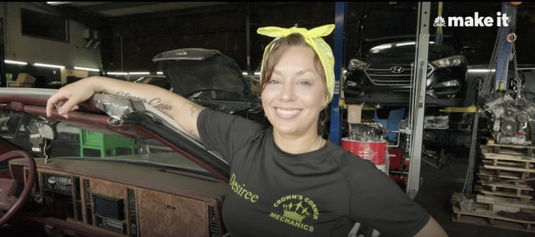 Mom proudly standing and smiling in her auto mechanic shop, in a black company t-shirt and a lemon yellow bandanna tied in a bow on top of her head.