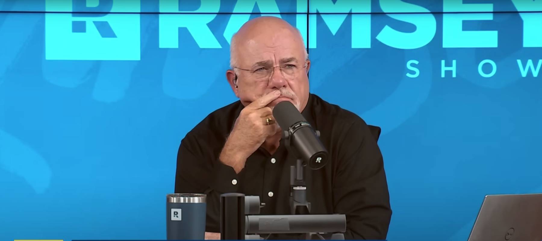 Dave Ramsey speaks into his microphone on set of the Ramsey Show, listening with his pointer finger on his lips in a thoughtful look.