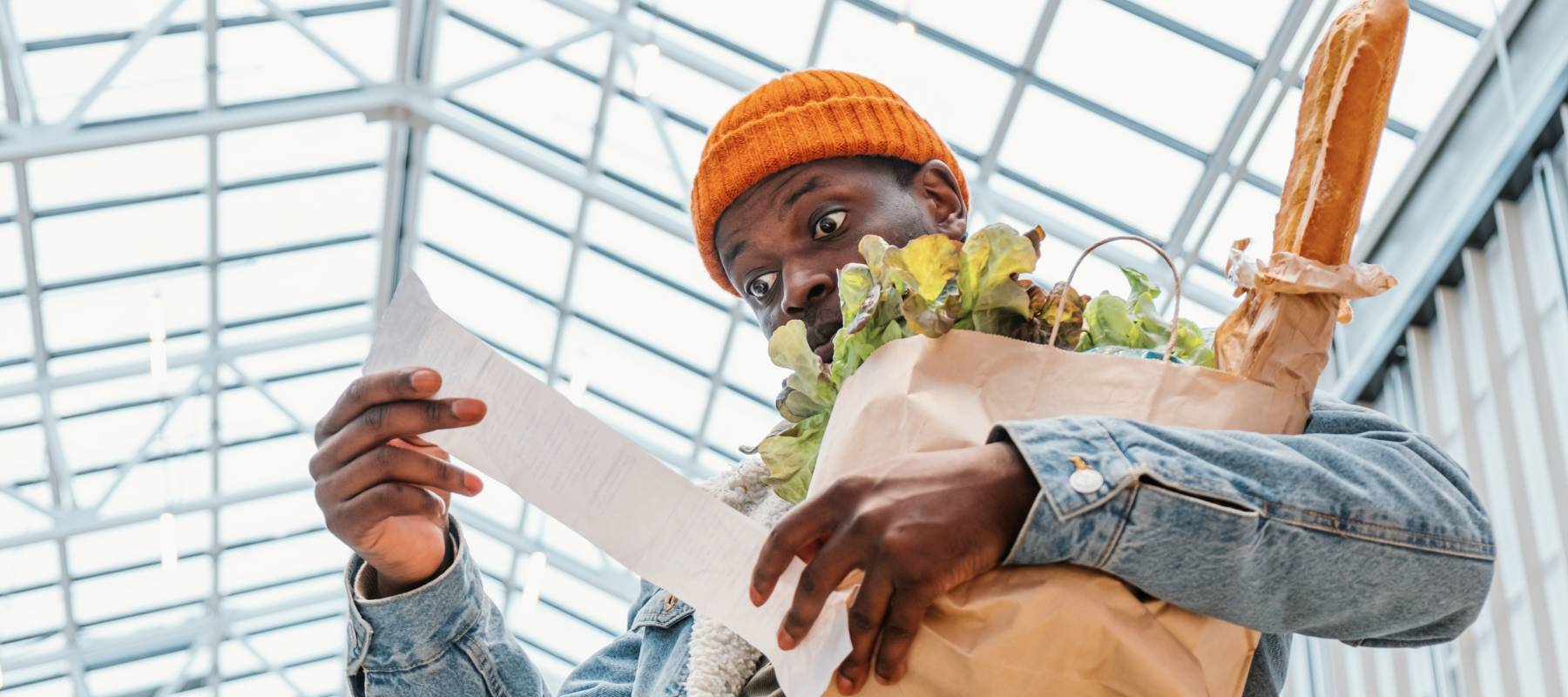 Surprised African-American man in denim jacket looks at receipt total in sales check holding paper bag with products in mall