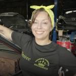 Mom proudly standing and smiling in her auto mechanic shop, in a black company t-shirt and a lemon yellow bandanna tied in a bow on top of her head.