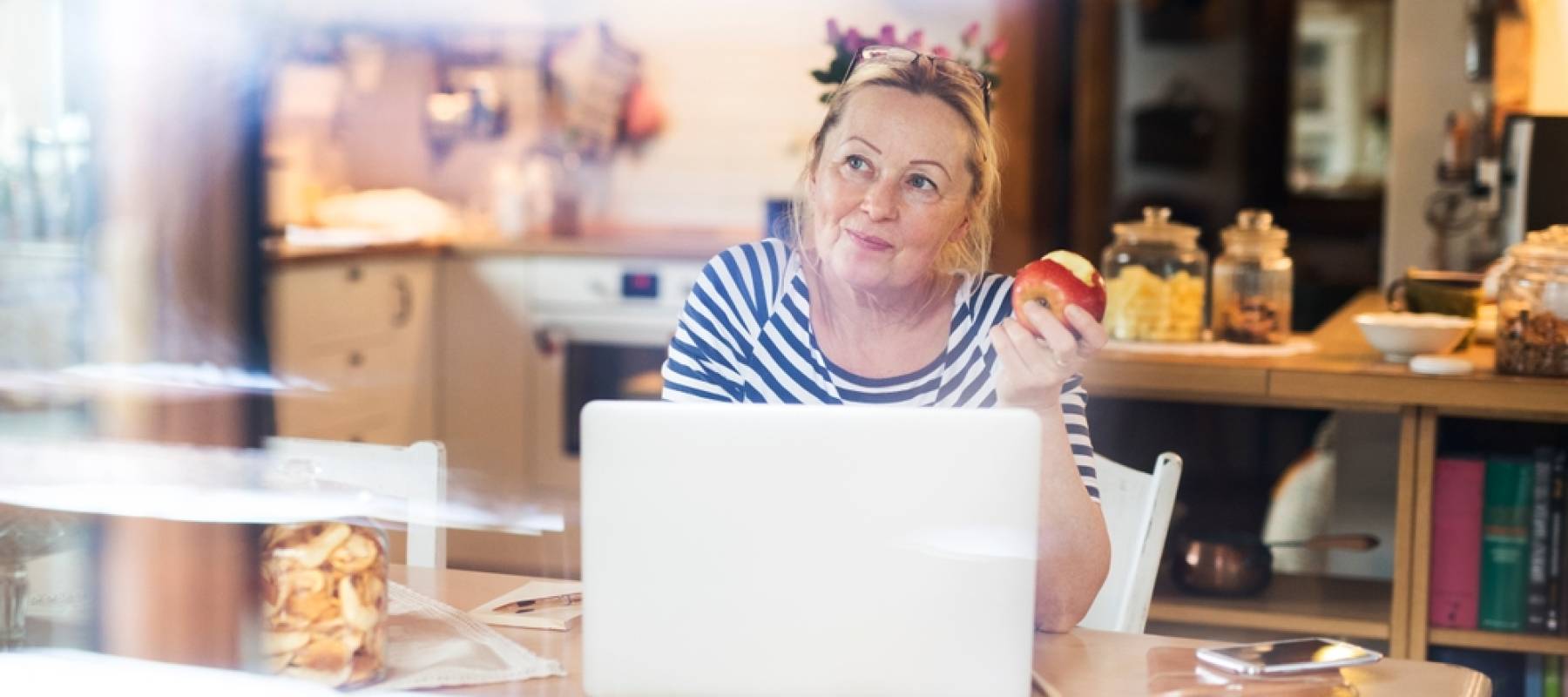 Older woman working on laptop, holding an apple in one hand