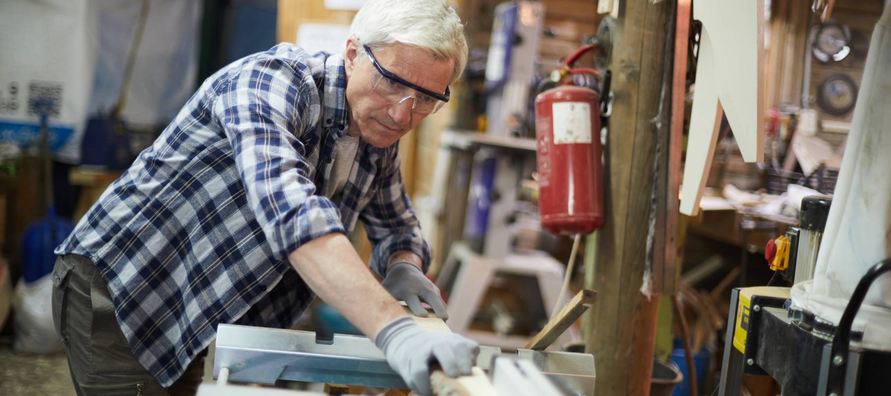 Aged master in protective eyeglasses processing wooden planks on machine in his workshop
