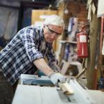 Aged master in protective eyeglasses processing wooden planks on machine in his workshop