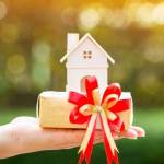 Woman hand holding a home model and gift box tied with red ribbon in the public park, for buying a new house or a real estate for family or person one loved concept.