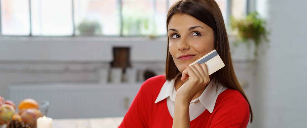 Undecided young woman making a purchase online sitting at home at her laptop holding her bank card in her hand and looking up with a thoughtful expression and wry smile