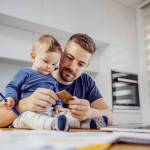 Young attractive bearded father sitting at dinning table with his beloved adorable son and paying bills online. His boy holding pen and trying to help him.