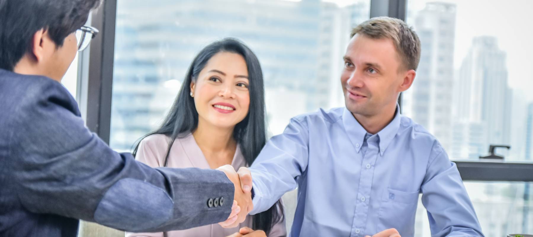 Couple smiling sitting in an office across a table from a man in a suit, men shaking hands.