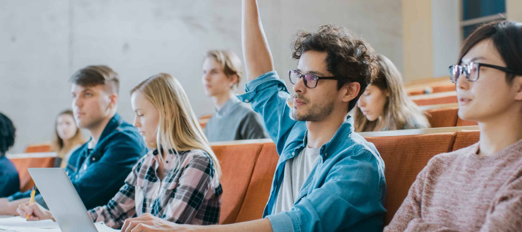 Handsome Hispanic Student Uses Laptop while Listening to a Lecture at the University, He Raises Hand and Asks Lecturer a Question. Multi Ethnic Group of Modern Bright Students.