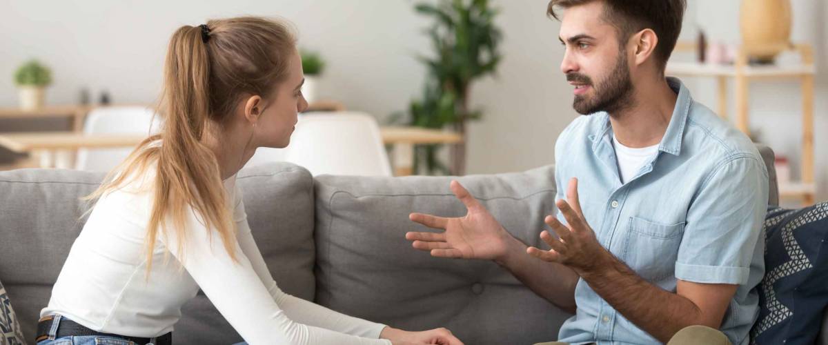 Serious young couple sitting together on sofa, talking about relationships, spending time together at home, focused wife listening to speaking husband, friends having conversation