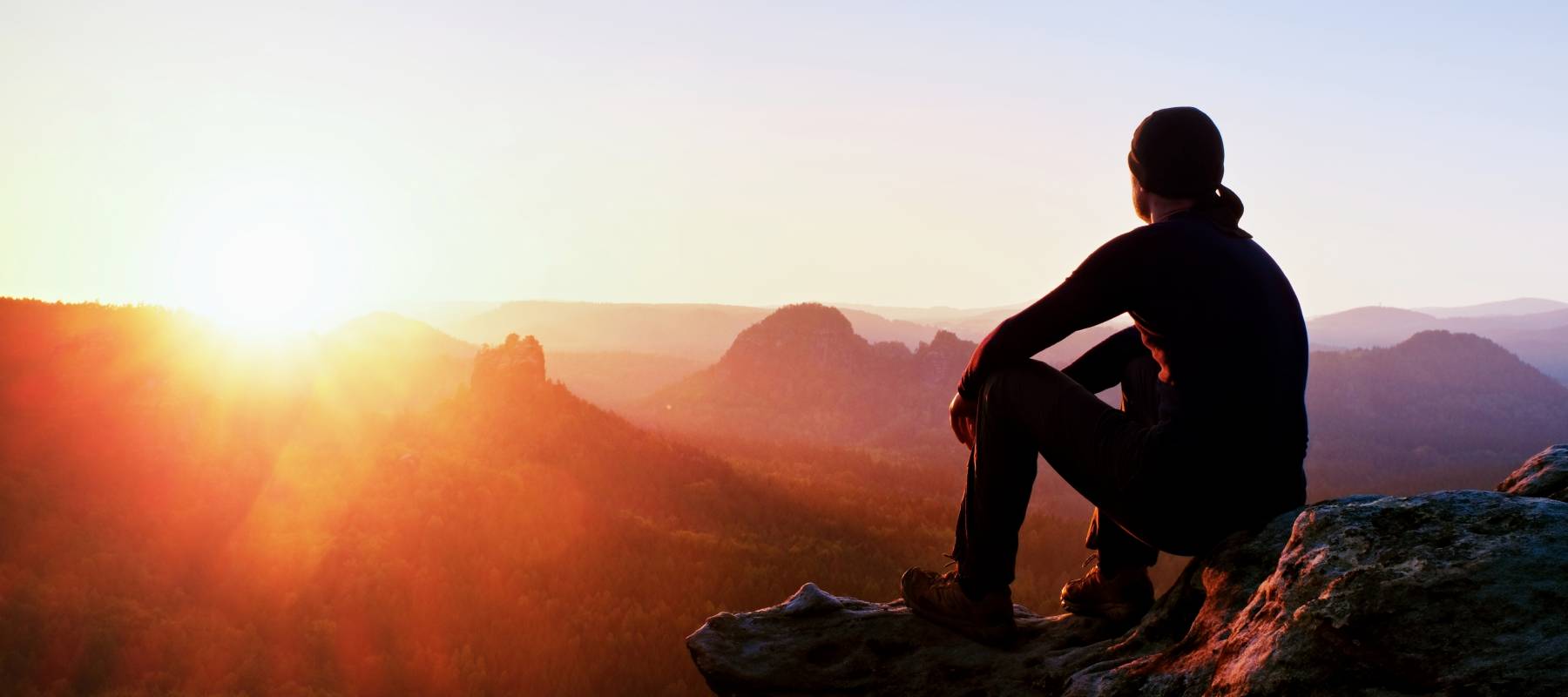 Adult tourist in black trousers, jacket and dark cap sit on cliff's edge and looking to misty hilly valley bellow