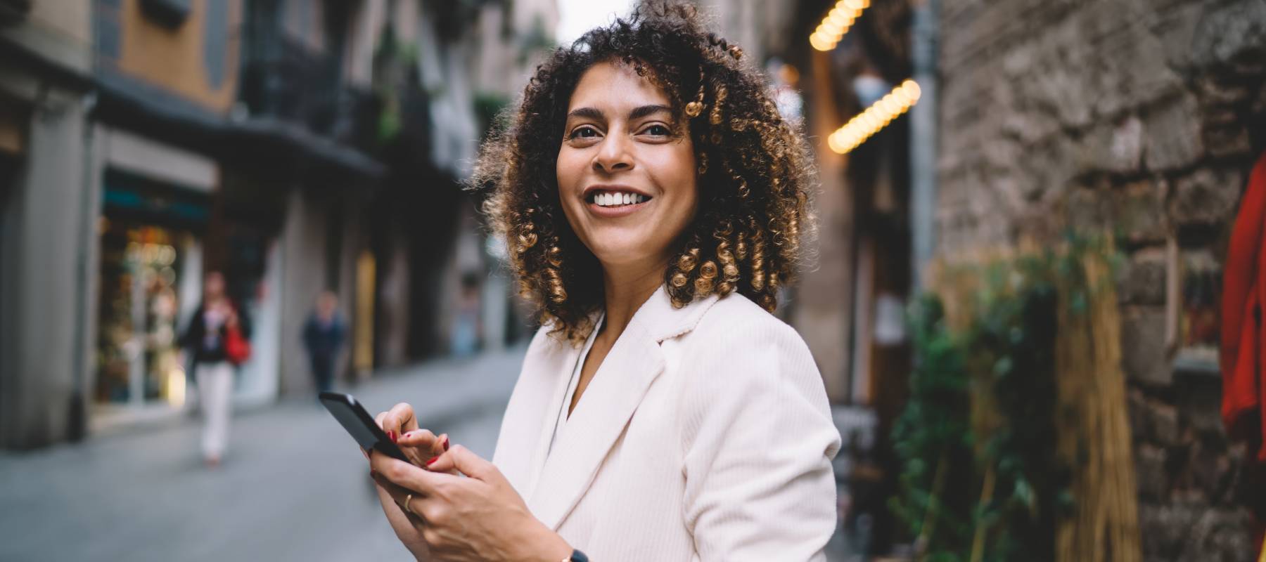 Young ethnic woman with smartphone smiling and standing on street and browsing internet wearing casual clothes on sunny day looking forward.