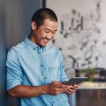 Casually dressed young Asian designer smiling and using a digital tablet while leaning against a gray wall in a modern office
