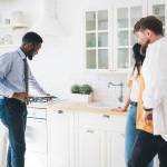 Side view of joyful business African American realtor presenting new kitchen to young buyers in big light flat and touching cookstove.