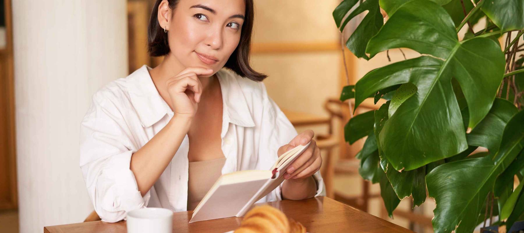 Smiling beautiful girl with a book, reading with thoughtful smirk, thinking of something, drinking coffee in cafe.