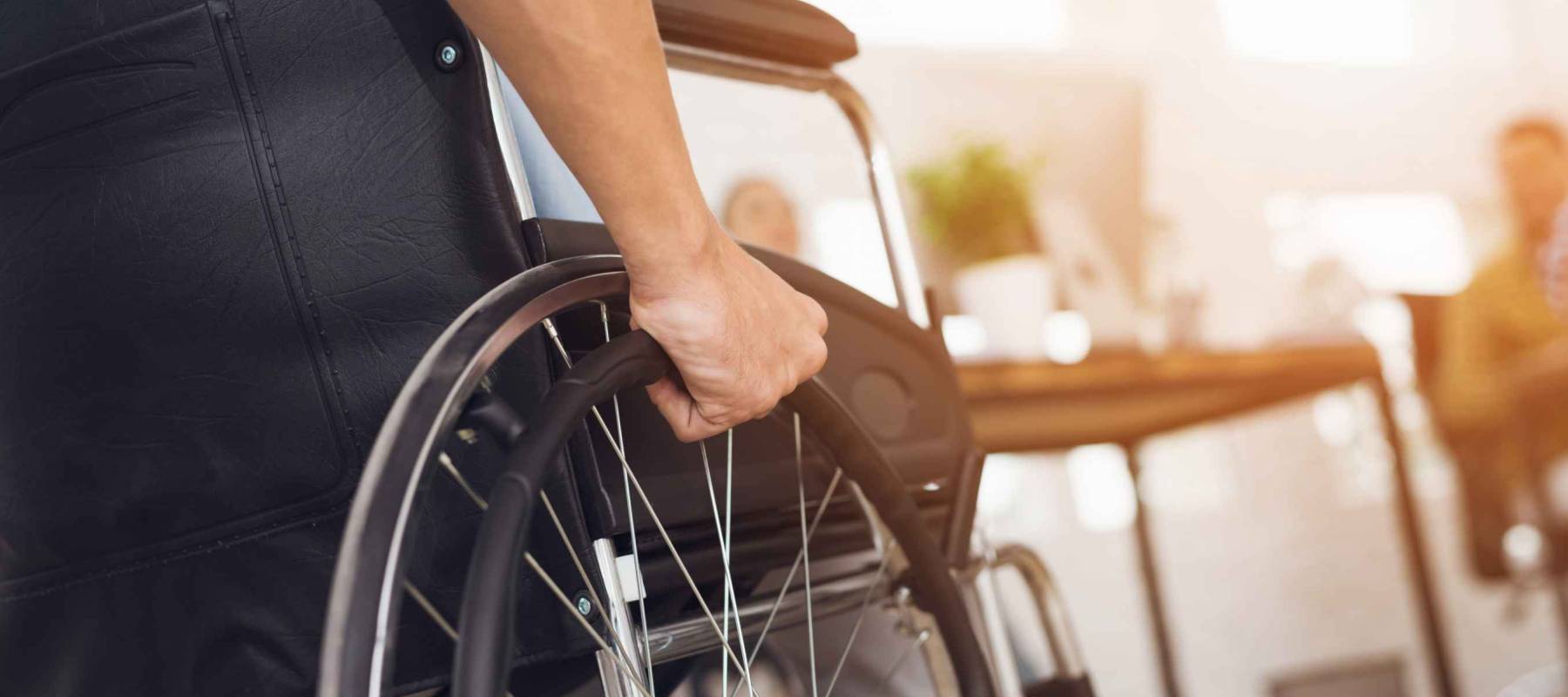 A disabled man is sitting in a wheelchair. He holds his hands on the wheel. Nearby are his colleagues