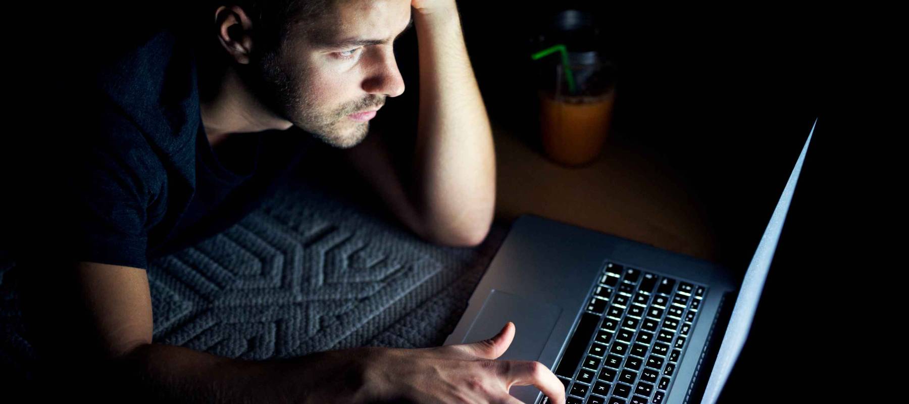 Top view of young man reading news on laptop lying in dark room. Looking concentrated