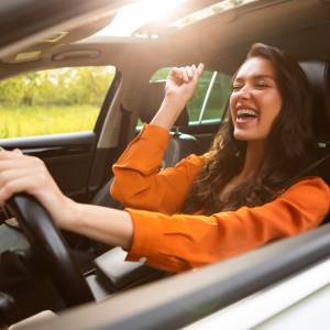 Overjoyed pretty young woman driver listening to music and singing while holding one hand on steering wheel, enjoying auto ride