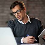 Multi-tasking businessman working in the office. He is using touchpad while reading an e-mail on laptop and taking notes on the paper.