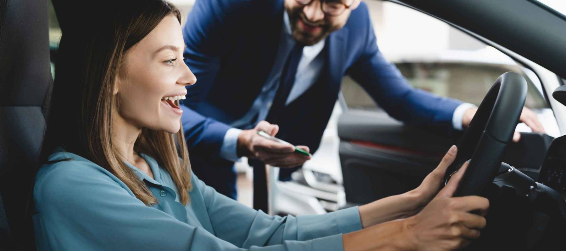 Satisfied happy caucasian female client customer woman sitting at the wheel of new car doing test-drive before buying auto while male shop assistant helping her choose it