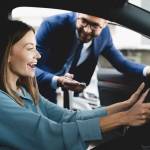 Satisfied happy caucasian female client customer woman sitting at the wheel of new car doing test-drive before buying auto while male shop assistant helping her choose it