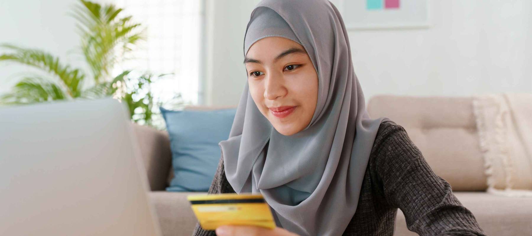 Muslim young women wear hijab are buying online with a credit card while sitting living room at home. Beautiful woman is using computer laptop and doing online transactions