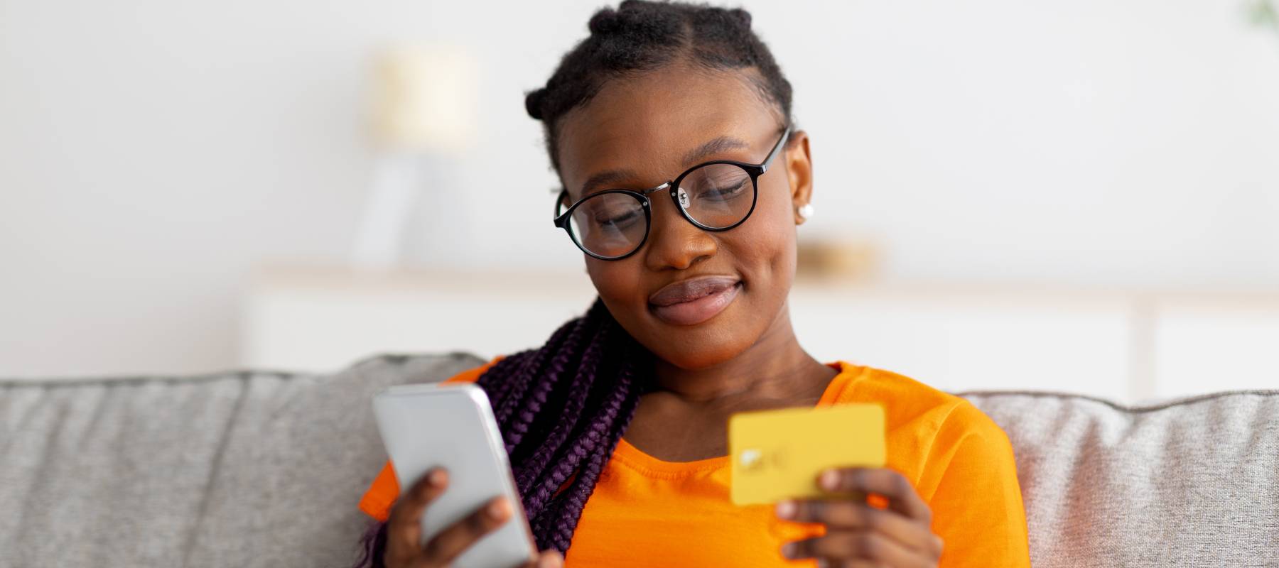 Lovely young black woman sitting on sofa, using mobile phone and credit card