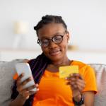 Lovely young black woman sitting on sofa, using mobile phone and credit card