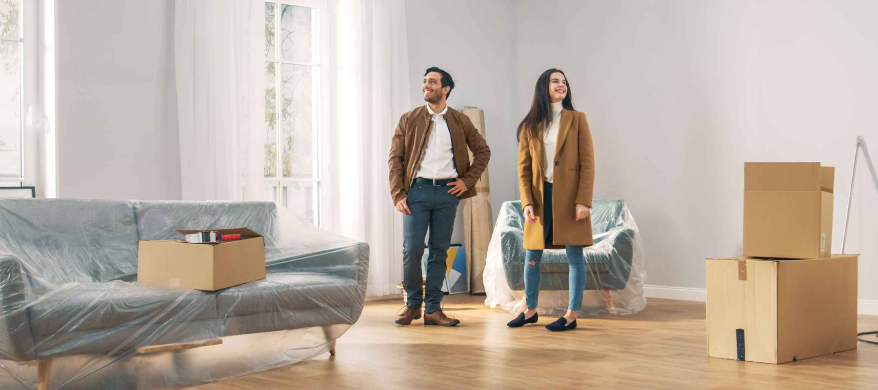 Happy and Excited Young Couple Look Around In Wonder at their Newly Purchased / Rented Apartment. Beautiful People Poses Happily. Big Bright Modern Home with Cardboard Boxes Ready to Unpack.
