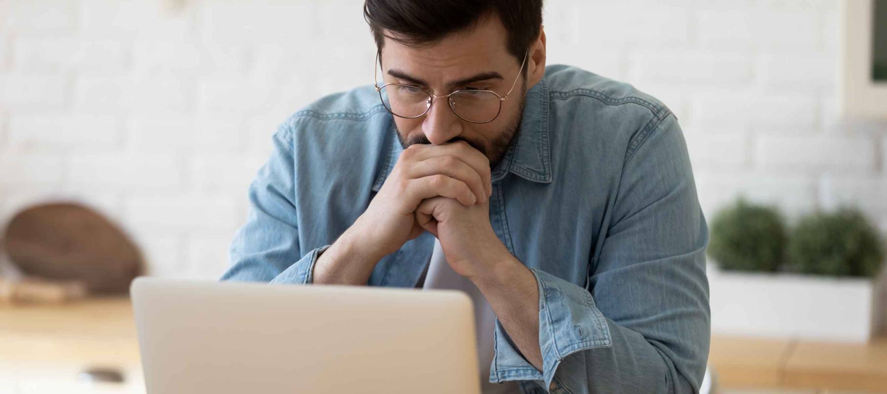 Head shot thoughtful businessman in eyewear looking at computer screen, sitting at table at home. thinking of problem solution