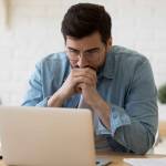 Head shot thoughtful businessman in eyewear looking at computer screen, sitting at table at home. thinking of problem solution
