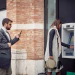 Man looking at phone waits in line to use an ATM as a woman takes money out of her bank account