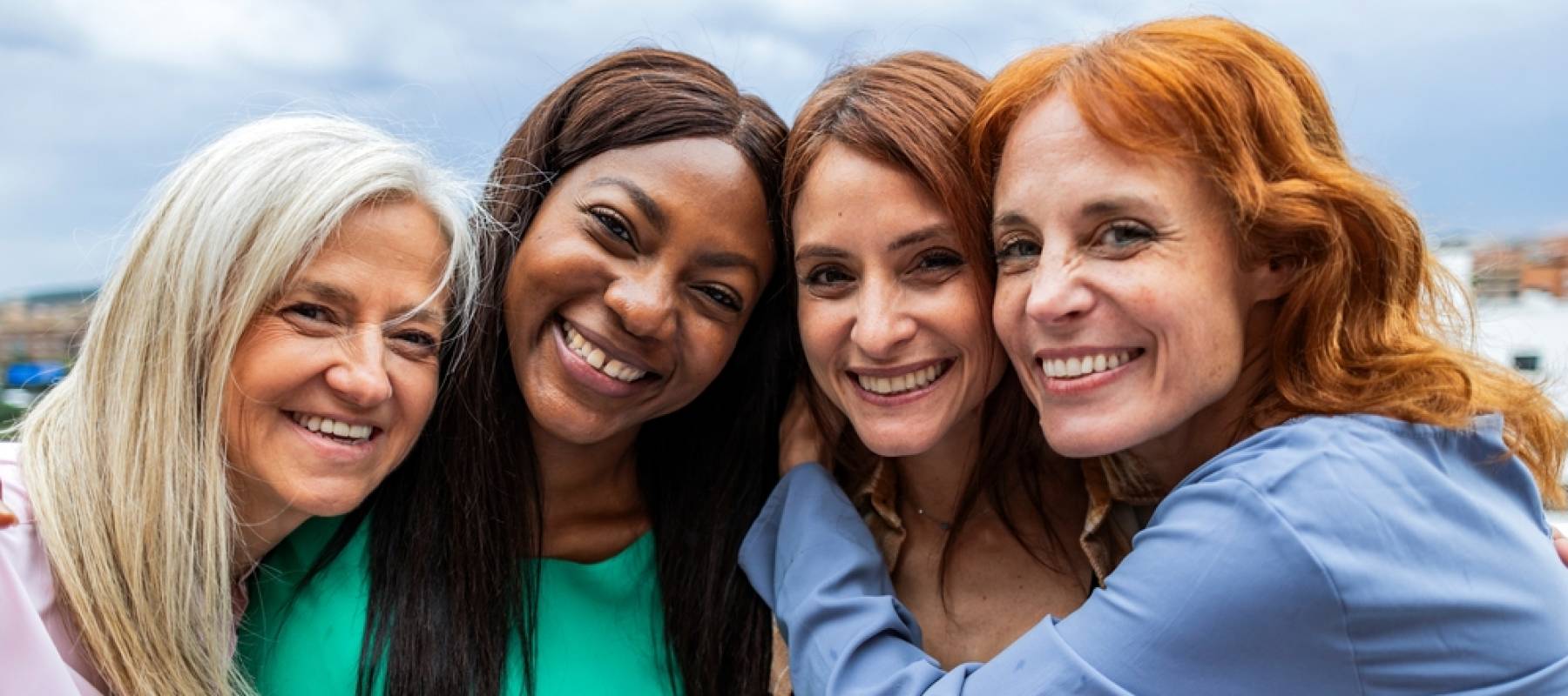 Four vibrant women huddled in a group hug, smiling brightly, with the open sky behind them.