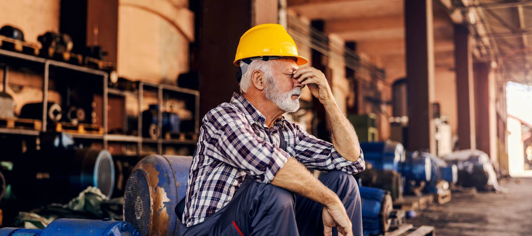 Senior factory worker is sitting next to the machines and holding his head