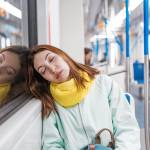 A brightly-dressed Gen Z female riding the subway asleep, and resting her head against the window.