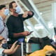 Family in face masks looking at their flight times on board at airport. Tourist man with wife and kid waiting at airport with luggage on trolley during pandemic.