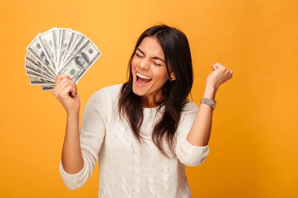 Portrait of a cheerful young woman holding money banknotes and celebrating isolated over yellow background