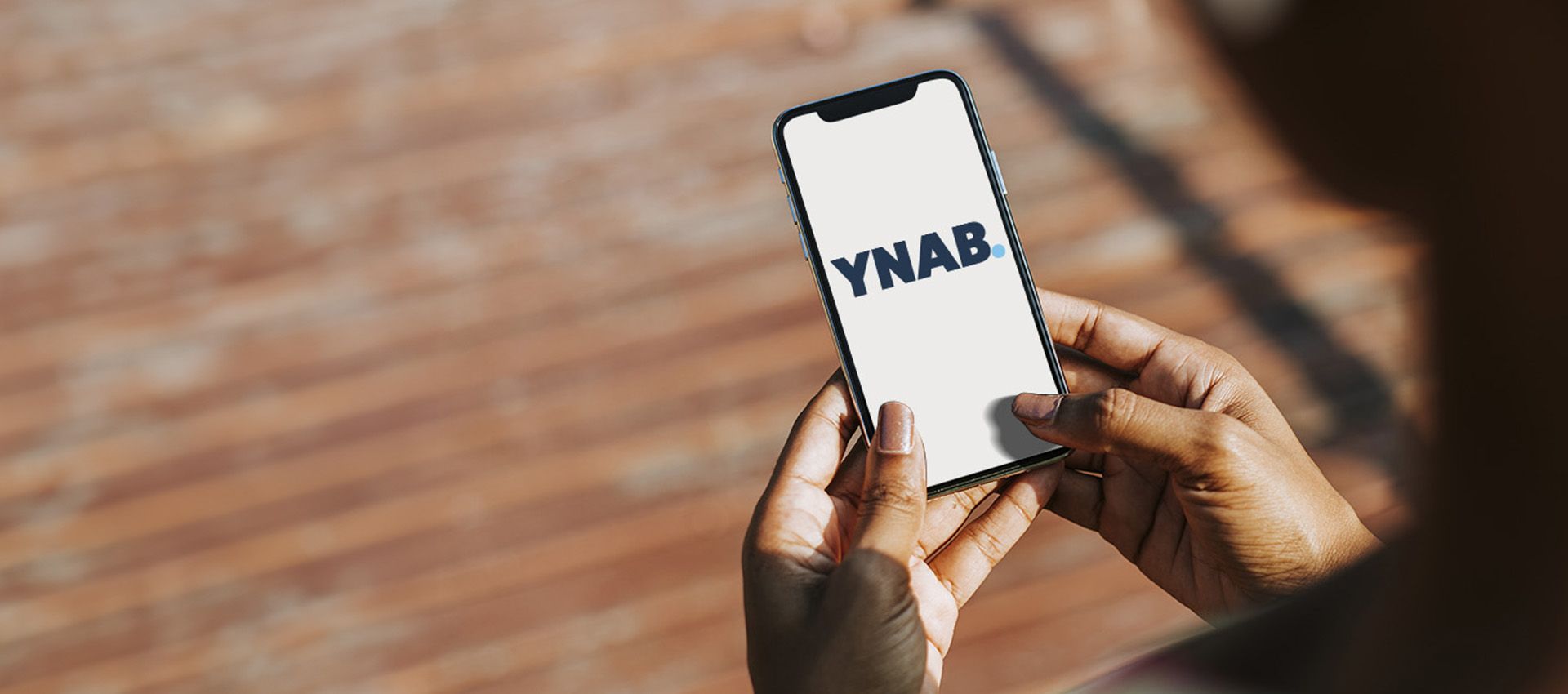 YNAB logo on mobile device being held in the hands of black woman with interlock brick ground in the background