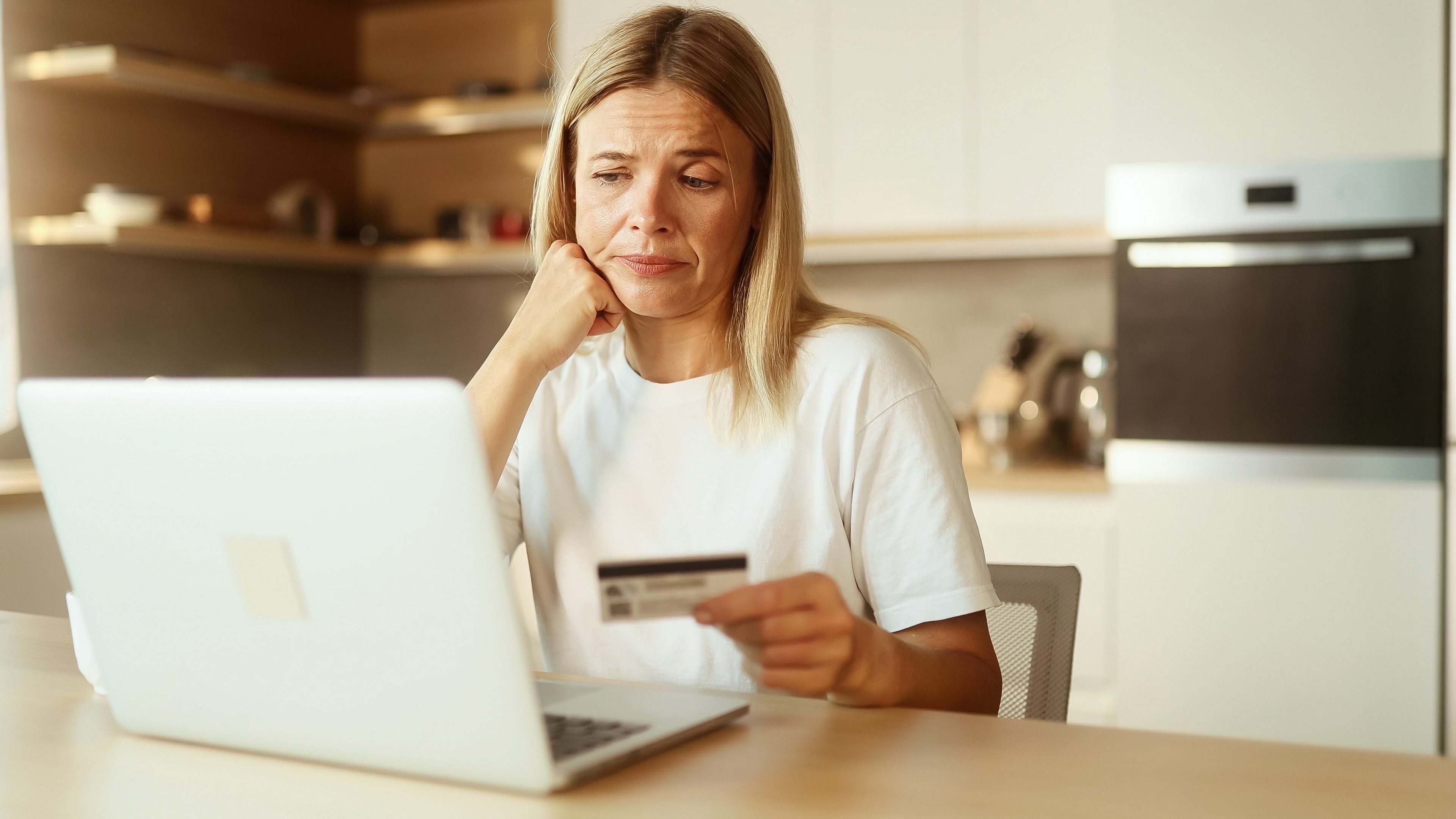 Confused blond woman with credit card thoughtfully pondering on screen while has problem with online order payment error or blocked bank account doing shopping at internet on laptop at light home