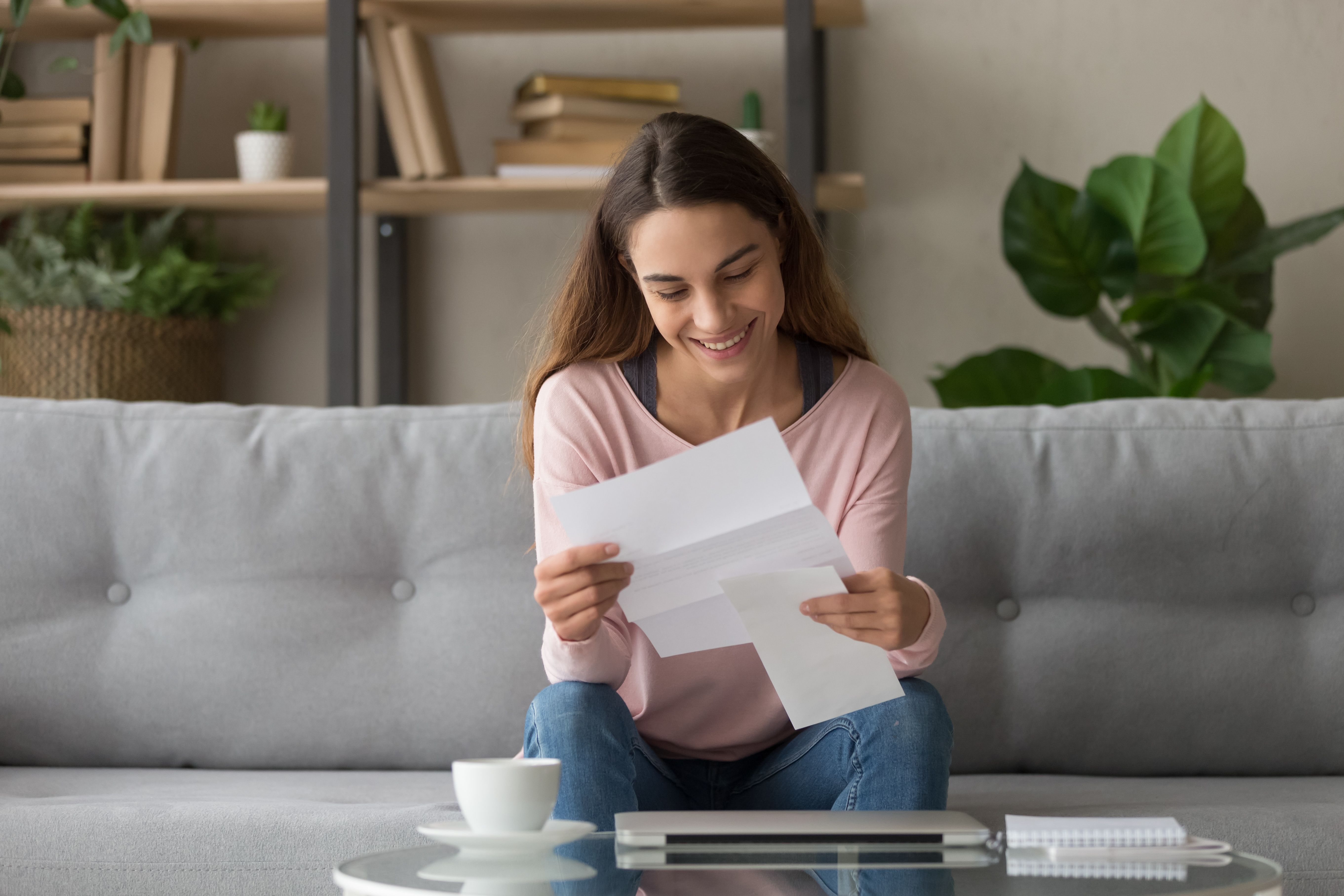 Happy smiling millennial girl holding paper document, received good news letter, university admission notification, last banking debt payment, high paid job offer, sitting on comfortable sofa at home.