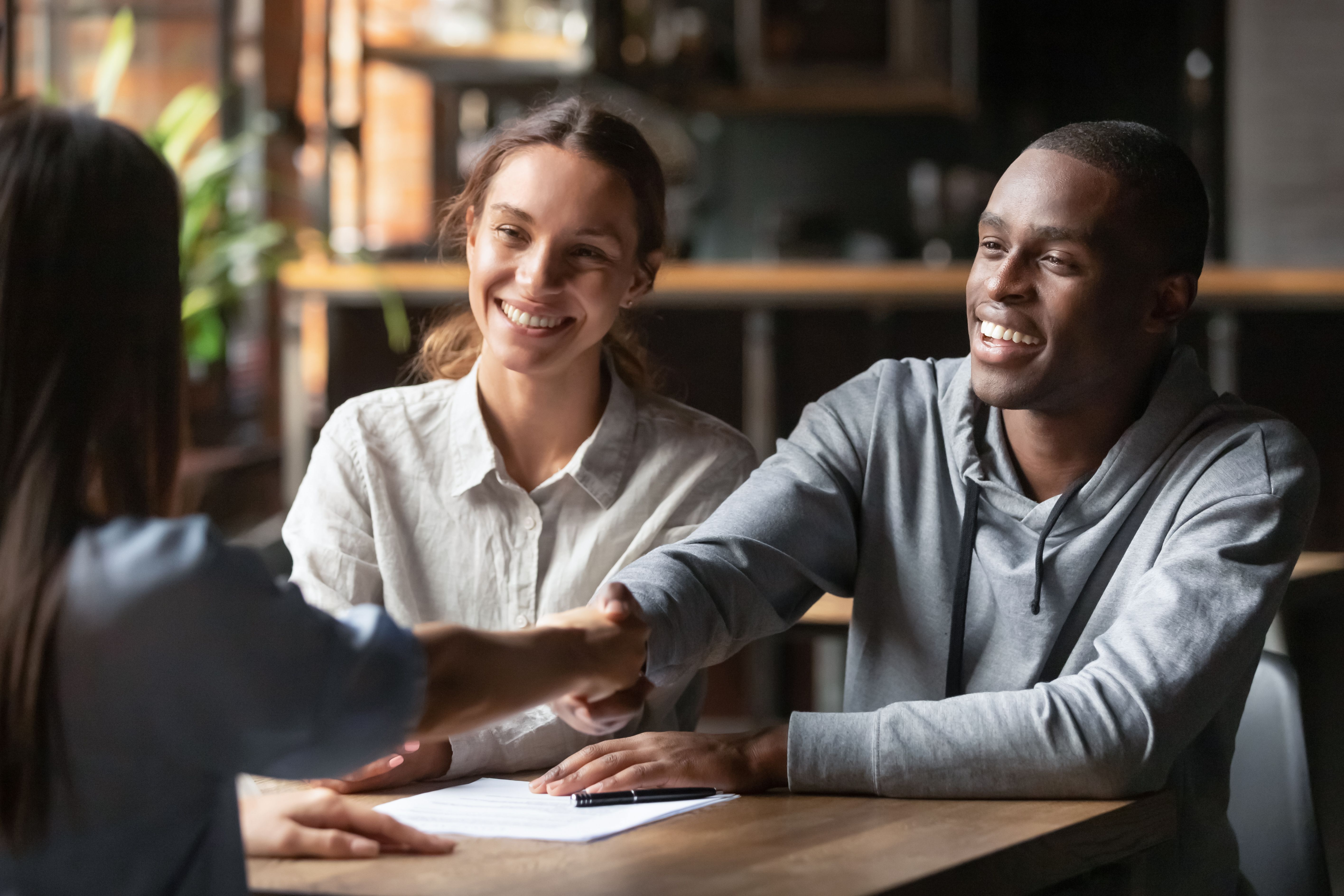Happy young interracial couple shake hand of bank manager broker buy insurance services take mortgage loan, mixed ethnicity customers handshake agent make agreement financial business deal at meeting