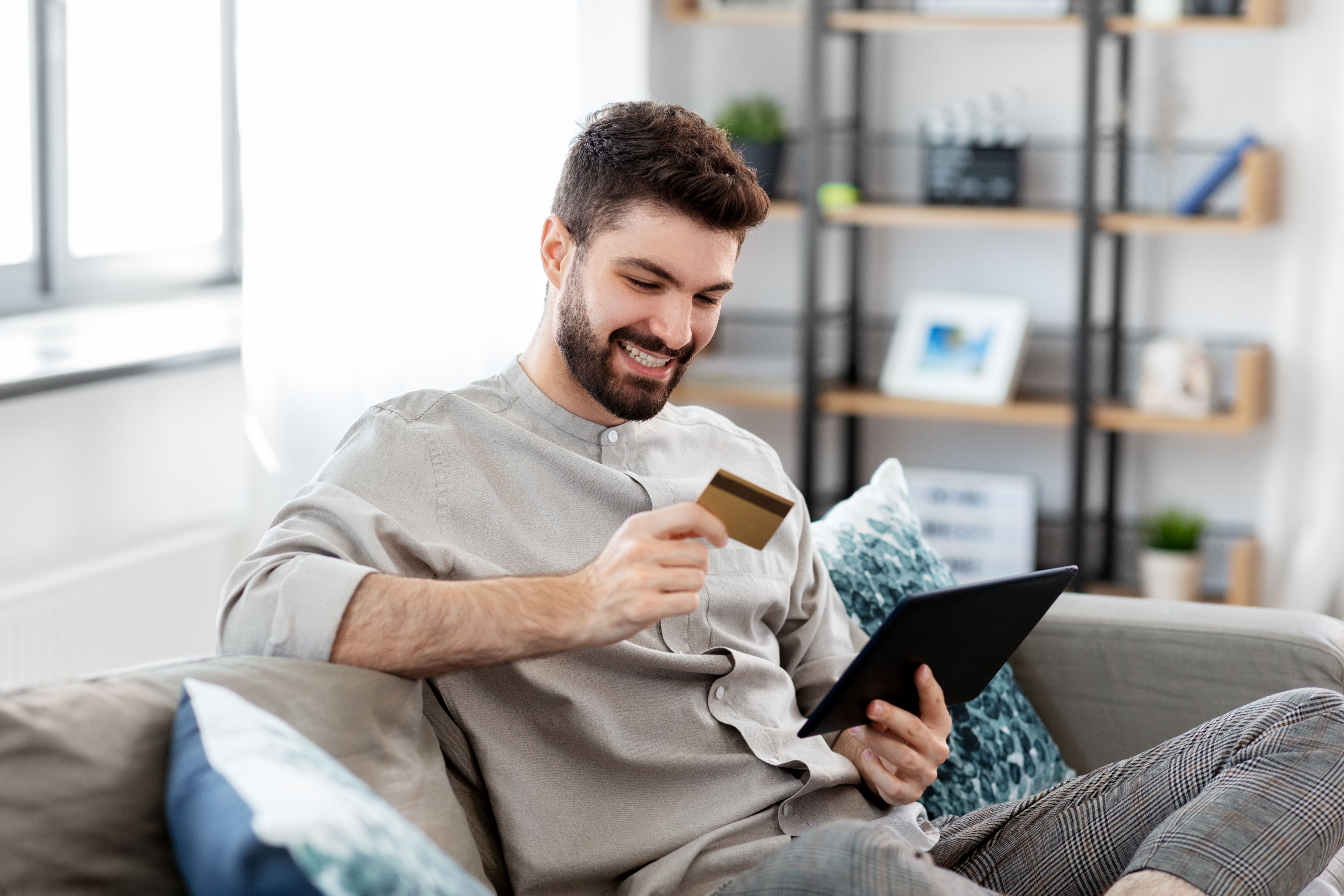 Handsome young smiling man with credit card and phone in hands doing purchases during online shopping