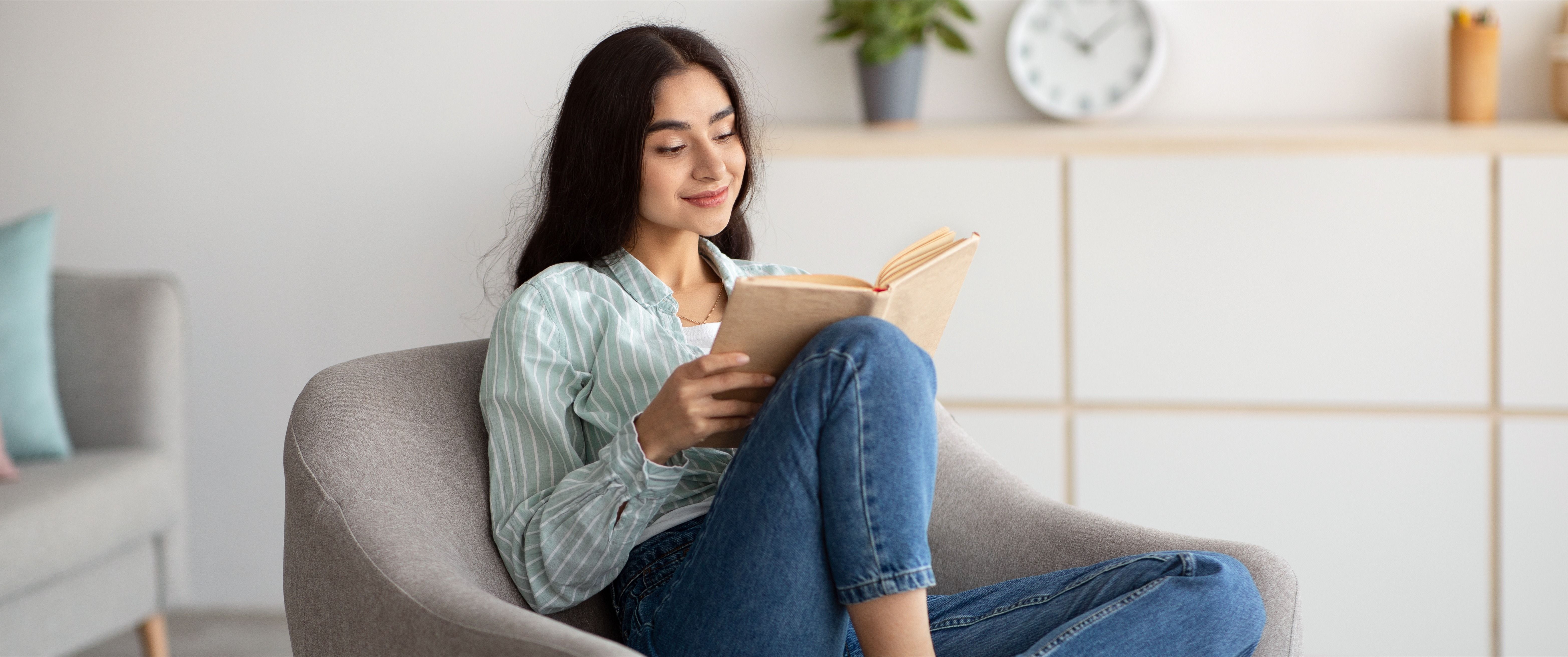 Millennial Indian woman sitting in cozy armchair with open book indoors.