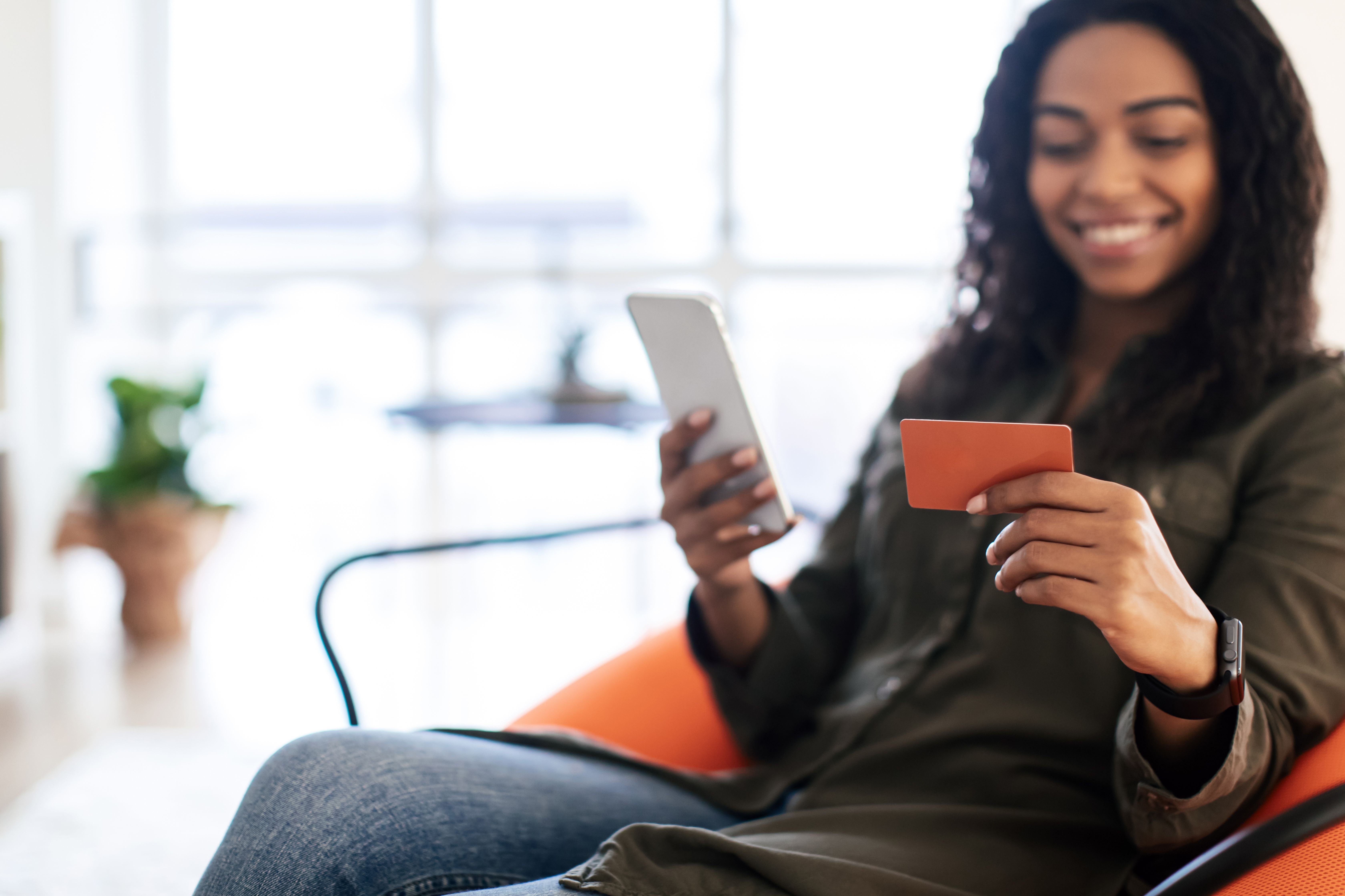 Fast Online Shopping. Smiling young black lady holding debit credit card and using cell phone, sitting on the bean bag pouf chair at home in living room, looking at bankcard in hand, free copy space