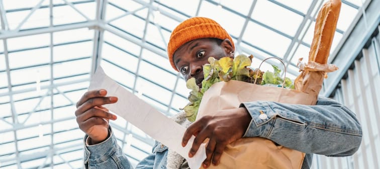 Surprised African-American man in denim jacket looks at receipt total in sales check holding paper bag with products in mall