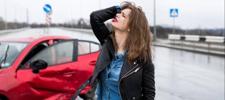 An upset young woman stands in front of a car that's been in a car accident