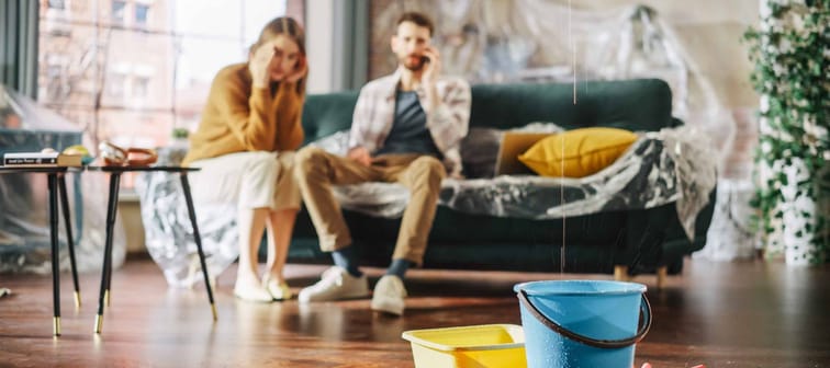 Roof is Leaking or Pipe Rupture at Home: Panicing Couple In Despair Sitting on a Sofa Watching How Water Drips into Buckets in their Living Room.